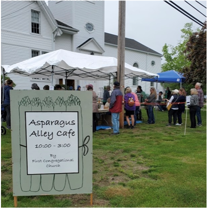 Image of a sign that says Asparagus Alley Cafe. Behind the sign is a tent in front of the church with people in line to buy asparagus chowder.